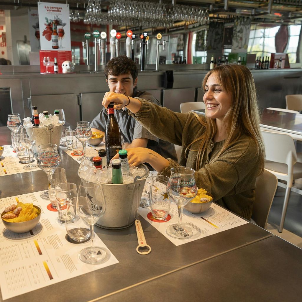 한 여성과 한 남성이 음료, 간식, 금속 얼음 양동이가 놓인 테이블에 앉아 캐주얼한 실내 분위기에서 미소를 짓고 있습니다.