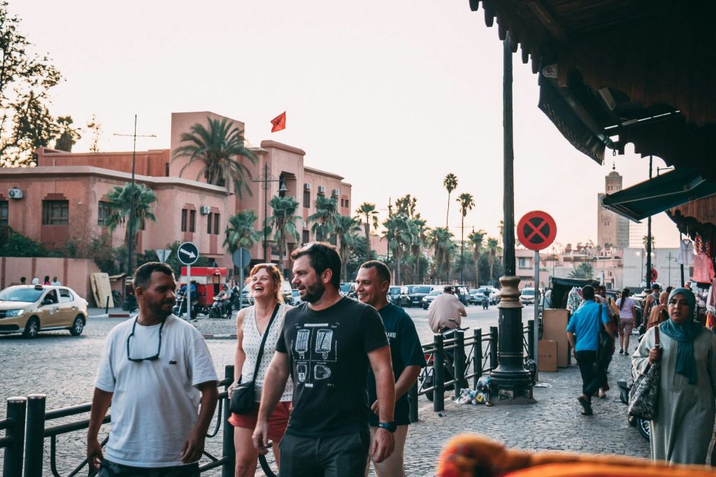 야자수가 우거진 도시 거리와 붉은 깃발을 배경으로 한 베이지색 건물에서 네 명이 함께 걸으며 웃고 있습니다.