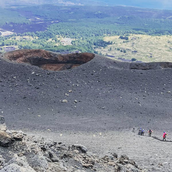 분화구를 배경으로 바위 화산 경사면을 오르는 등산객과 아래 녹색 풍경.