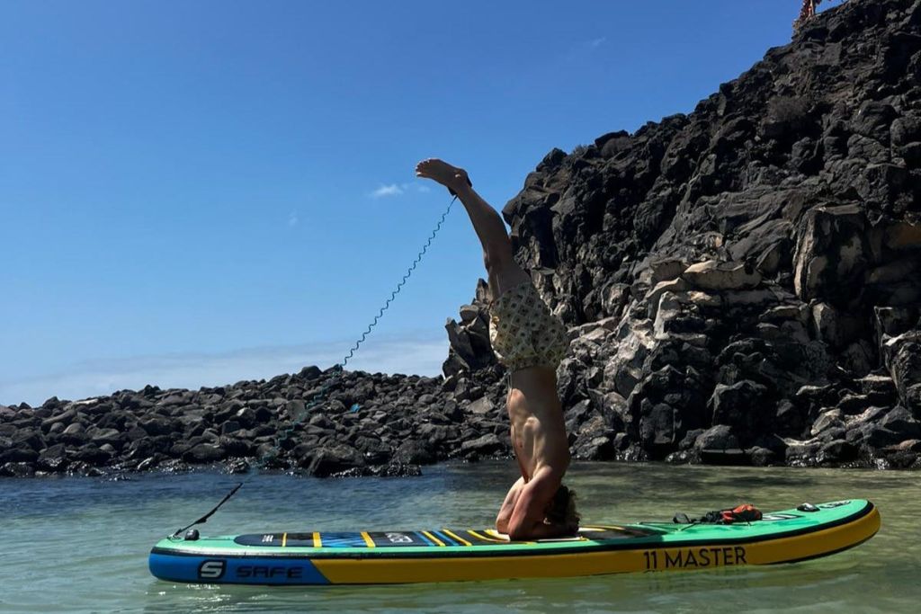 맑고 푸른 하늘 아래 바위 해안 근처의 잔잔한 물가에서 패들보드 위에서 헤드스탠드를 하고 있는 사람.