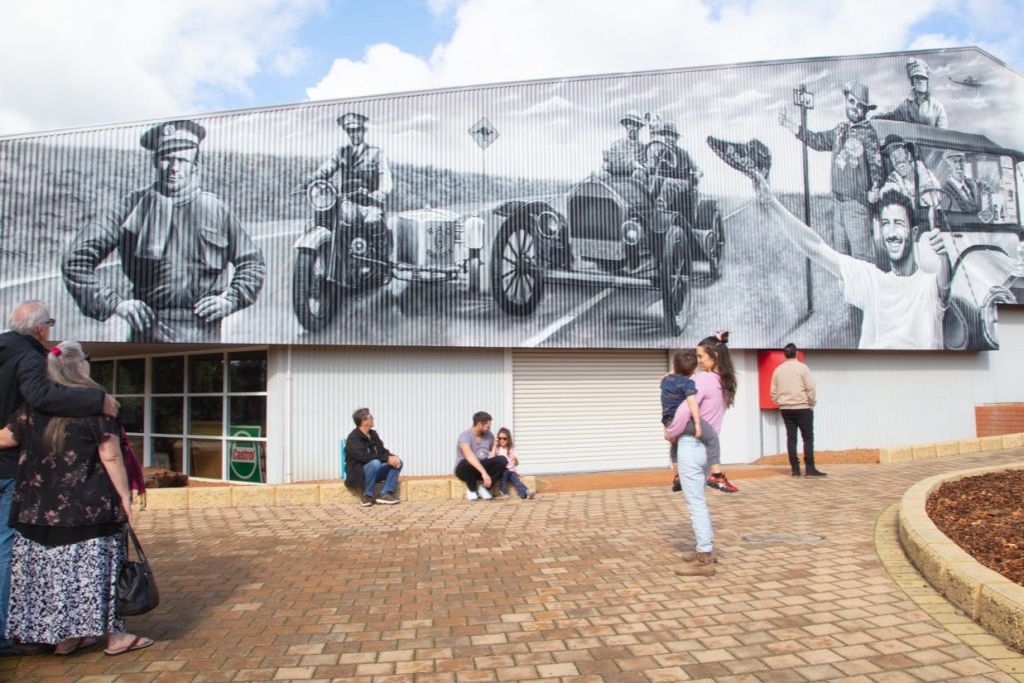 어린이를 안고 있는 한 여성이 사람과 빈티지 자동차를 묘사한 커다란 벽화가 있는 건물 앞에 서 있습니다. 여러 사람이 근처에 앉아 있습니다.