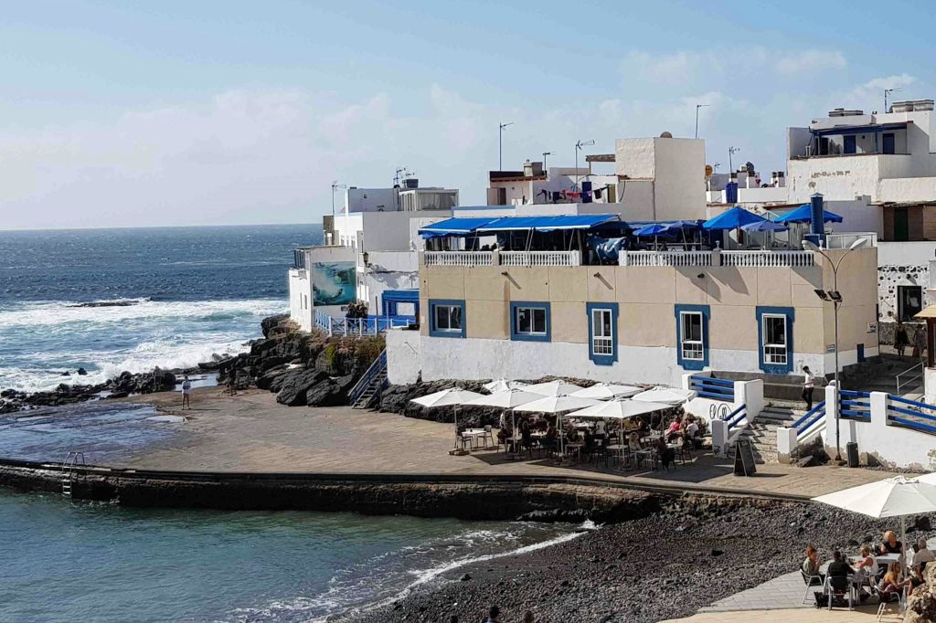 파란색 테두리가 있는 해변 건물, 파티오에 흰색 파라솔이 놓인 야외 카페, 바위 같은 해안선, 파도를 배경으로 한 공간.