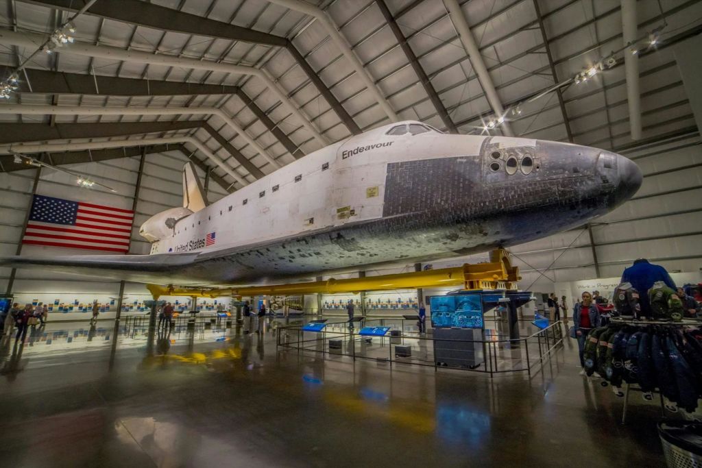 대형 박물관 격납고 내부에는 정보 전시물과 벽에 걸린 미국 국기로 둘러싸인 우주 왕복선 인데버 (Space Shuttle Endeavour) 가 전시되어 있습니다.