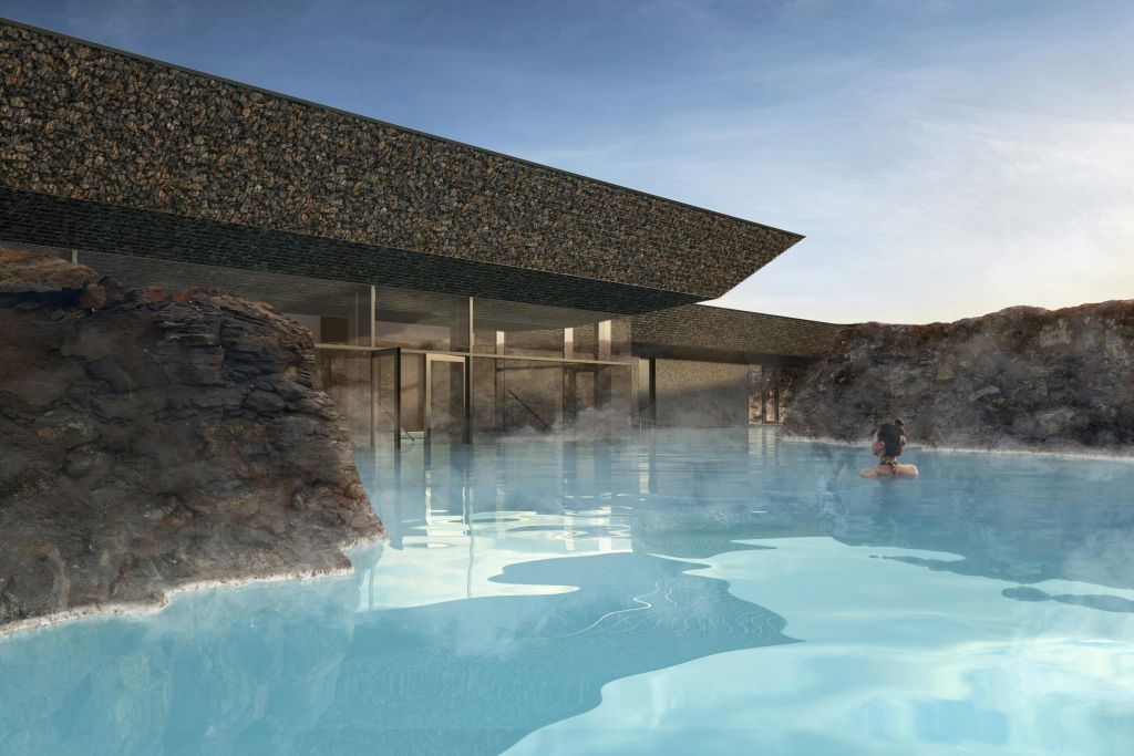 돌담이 있는 현대적인 건물은 바위로 둘러싸인 푸른 지열 수영장이 내려다보입니다. 한 사람이 수영을 하고 있습니다.