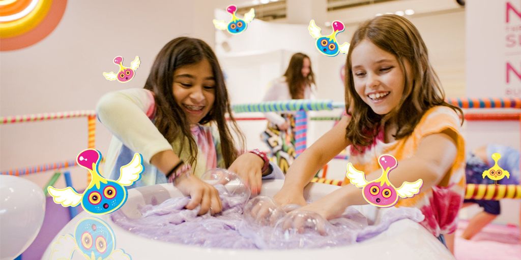 한 소녀가 다채로운 실내 놀이 공간에서 거품을 가지고 놀며 미소를 짓고 있습니다. 만화 날개 달린 생물들이 그녀를 에워싸고 있다.