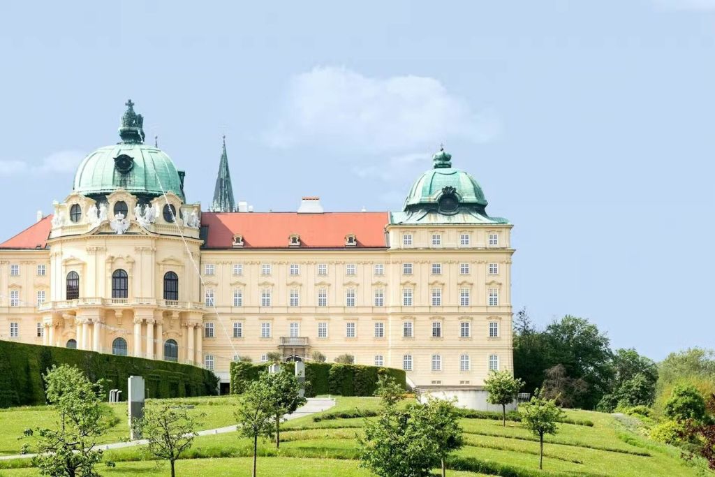 초록색 돔, 빨간색 지붕, 잘 다듬어진 정원이 있는 화려하고 밝은 베이지색 바로크 양식의 건물입니다. 구름이 거의 없는 푸른 하늘.