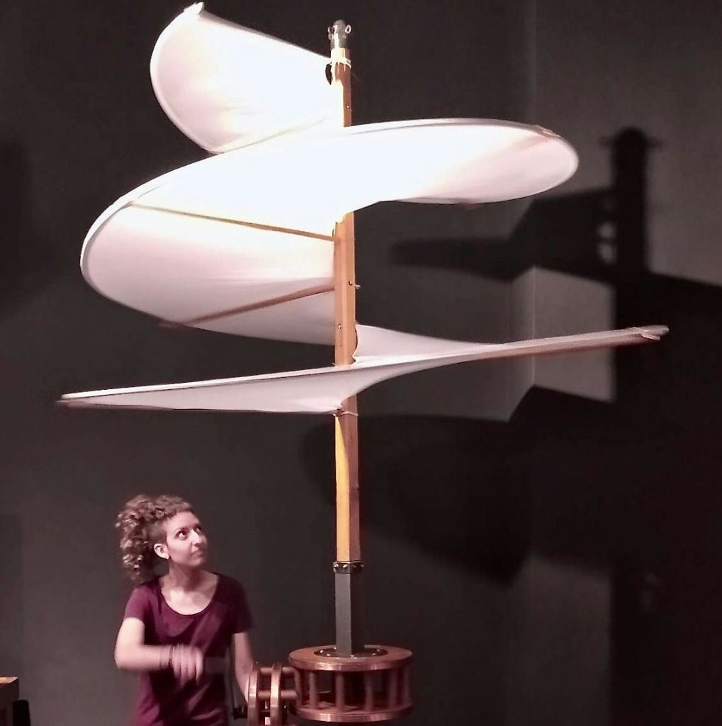 커다랗고 수직인 나선형의 날개 달린 구조물을 올려다보는 사람은 나무 받침이 있는 모형이나 조각품일 수 있습니다.