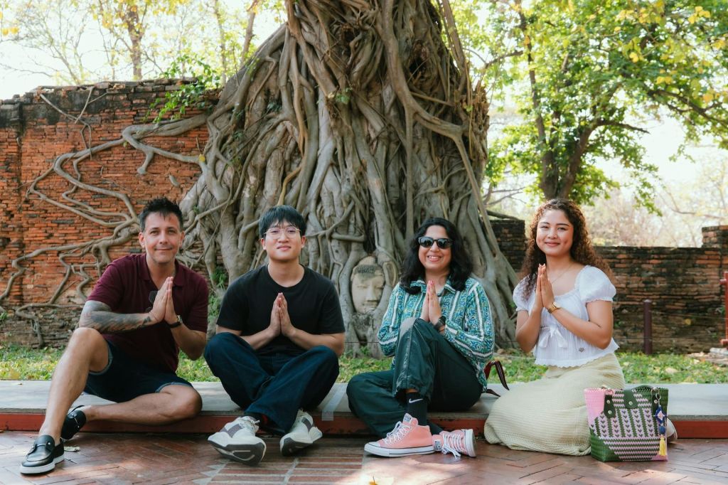 세 사람이 뿌리에 부처님 머리를 박은 채 나무 앞 바닥에 앉아 미소를 짓고 손바닥으로 포즈를 취하고 있습니다.