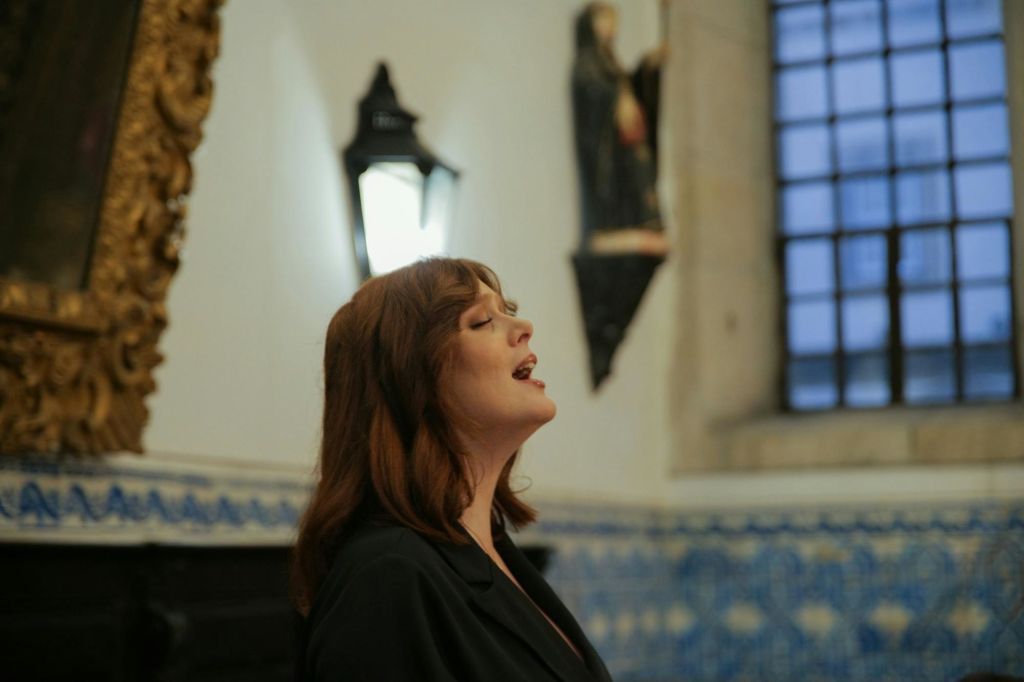 붉은 머리카락을 가진 사람이 조각상과 파란색 무늬의 타일 등 종교적인 장식이 있는 실내 환경에서 노래를 부르고 있습니다.