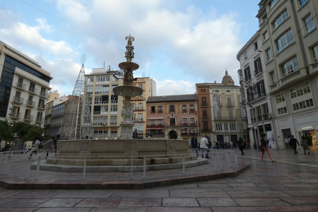 역사적이고 현대적인 건물, 걷는 사람들, 금속 프레임워크 구조로 둘러싸인 중앙 분수가 있는 광장입니다.