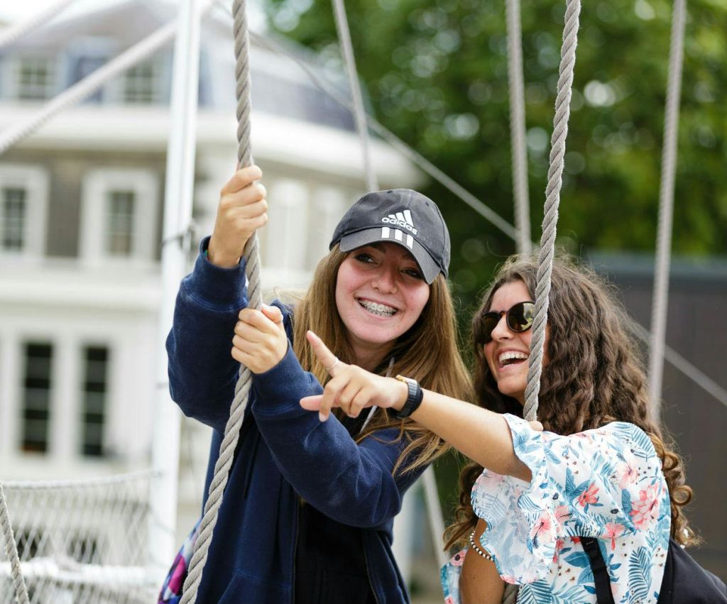 두 명의 미소 짓는 젊은 여성, 한 명은 중괄호가 달린 모자를 쓰고 다른 한 명은 선글라스를 쓰고 야외에서 밧줄을 잡고 캐주얼한 분위기를 연출합니다.