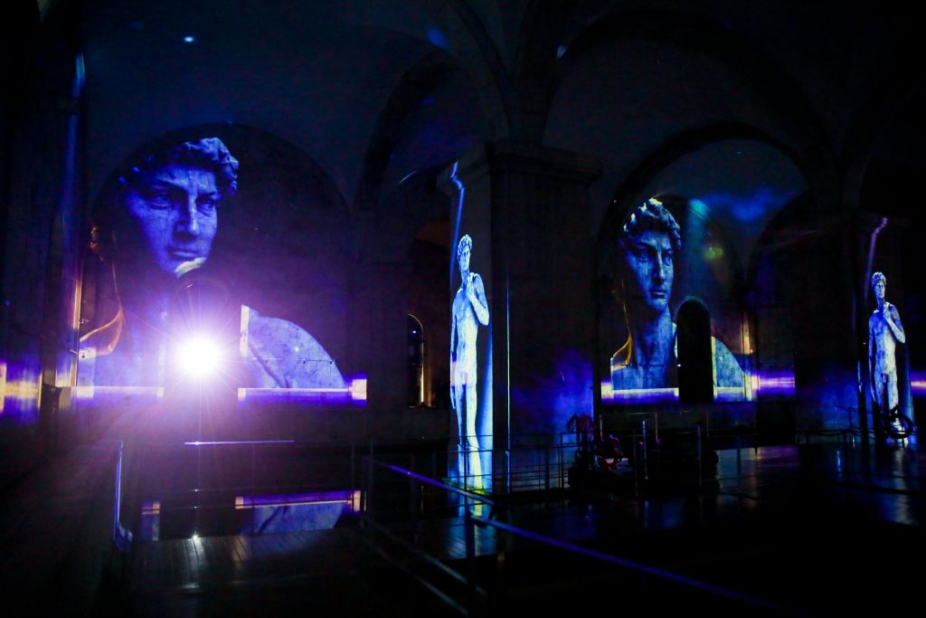 아치형 벽에 미켈란젤로의 다비드상이 투사된 커다란 이미지와 스포트라이트가 밝은 플레어를 연출하는 희미한 조명 공간.