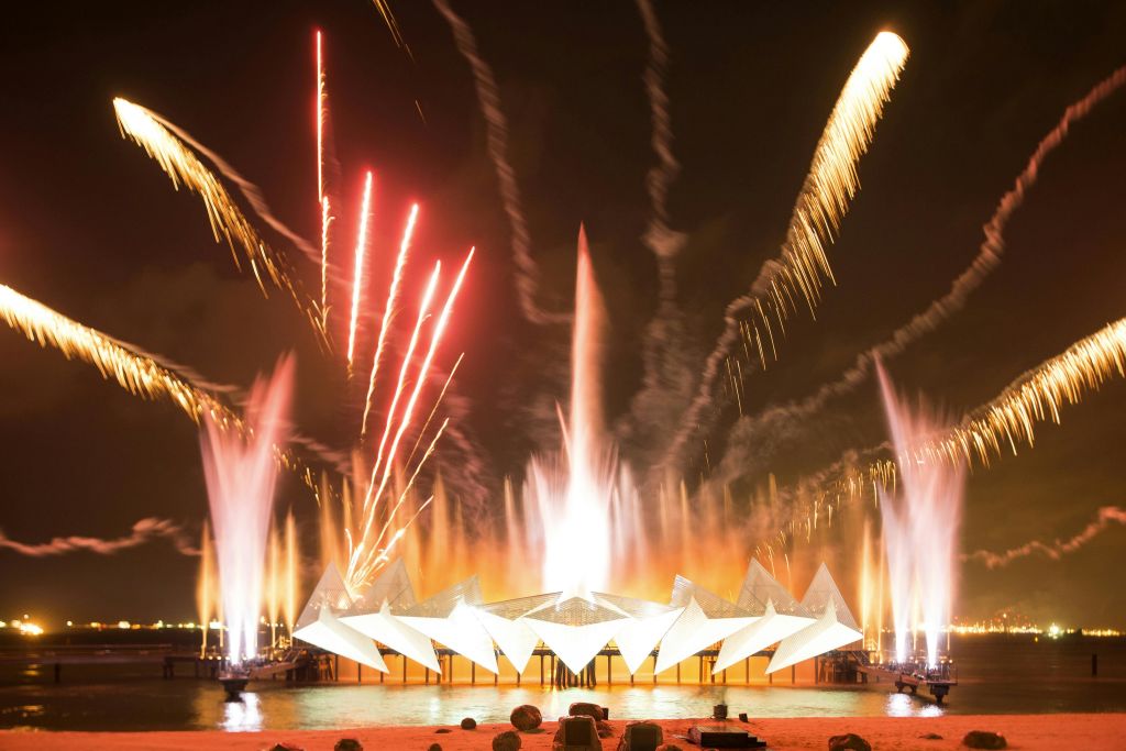 해안가의 기하학적 구조물 주변에는 밝은 불꽃놀이와 다채로운 분수가 밤하늘을 비추고 있습니다.