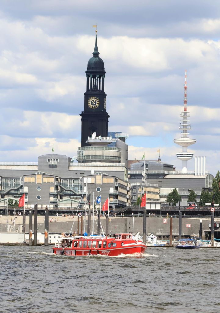 커다란 시계탑과 흐린 하늘 아래 현대적인 건물이 있는 붉은색 투어 보트가 도시 풍경을 배경으로 강을 항해합니다.