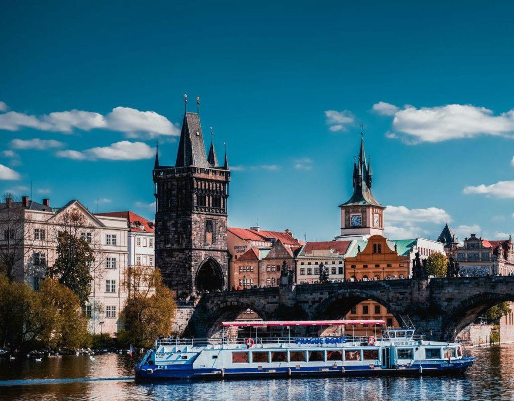 구름이 있는 푸른 하늘 아래 다리, 고딕 양식의 타워, 시계탑이 있는 유서 깊은 도시 풍경 앞에 있는 강 위의 보트.