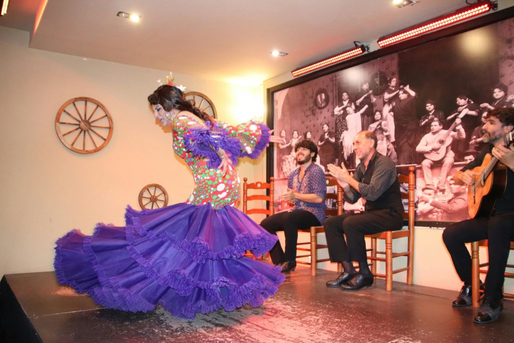 화려한 색상의 드레스를 입은 여성이 무대에서 춤을 추고 있으며, 남성이 무대 뒤에서 박수를 치고 있습니다. 장식에는 벽에 휠 장식이 포함됩니다.