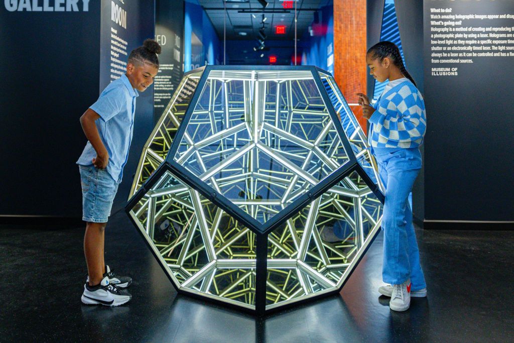 한 어린이가 현대적인 갤러리 공간에서 미러링된 표면이 있는 기하학적 조명 조형물을 살펴보고 있습니다.