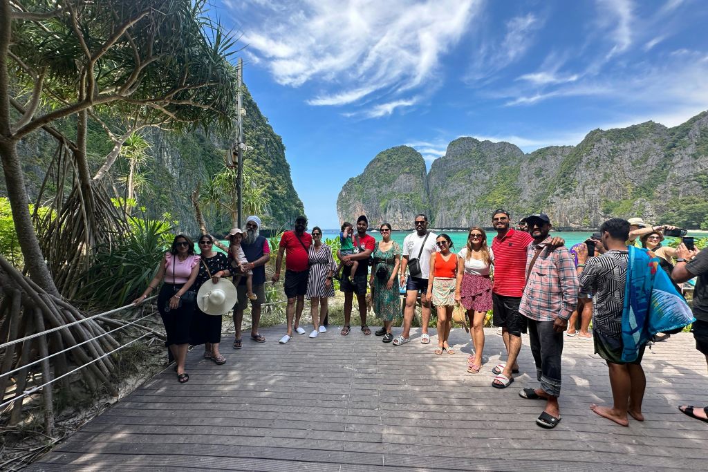 한 무리의 사람들이 절벽과 푸른 하늘을 배경으로 경치가 아름다운 나무 데크에 서서 미소를 짓고 사진을 찍으며 포즈를 취하고 있습니다.