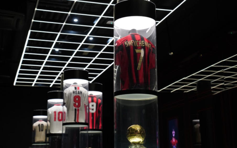 "Shevchenko 7" 과 "Weah 9" 를 포함한 원통형 디스플레이 케이스의 축구 유니폼. 황금색 축구공이 눈앞에 있습니다.