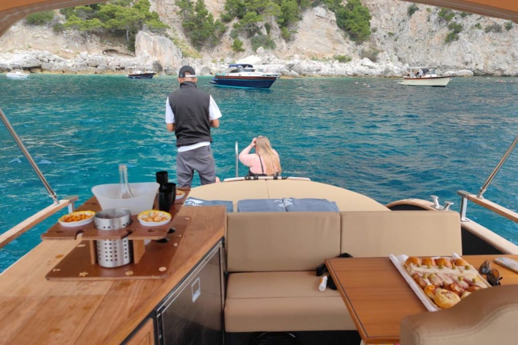 근처에 정박한 다른 보트와 함께 바위 해안 근처의 보트에 타고 있는 두 사람. 선상에는 테이블 위에 놓인 간식과 음료가 있습니다.