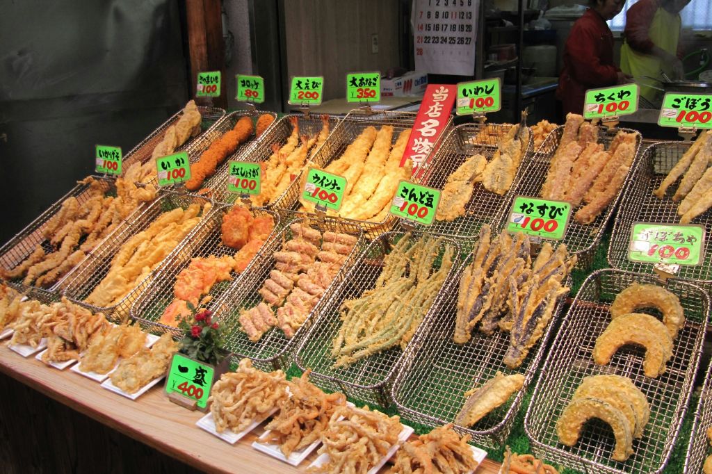 와이어 랙에 다양한 튀김 식품을 일본어로 다채로운 가격표와 함께 표시합니다. 시장이나 음식 가판대에 있을 가능성이 높습니다.