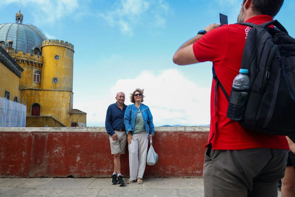 화창한 날 빨간색 셔츠를 입은 사람이 노란색 성탑 앞에서 사진을 찍기 위해 포즈를 취하고 있는 노부부.