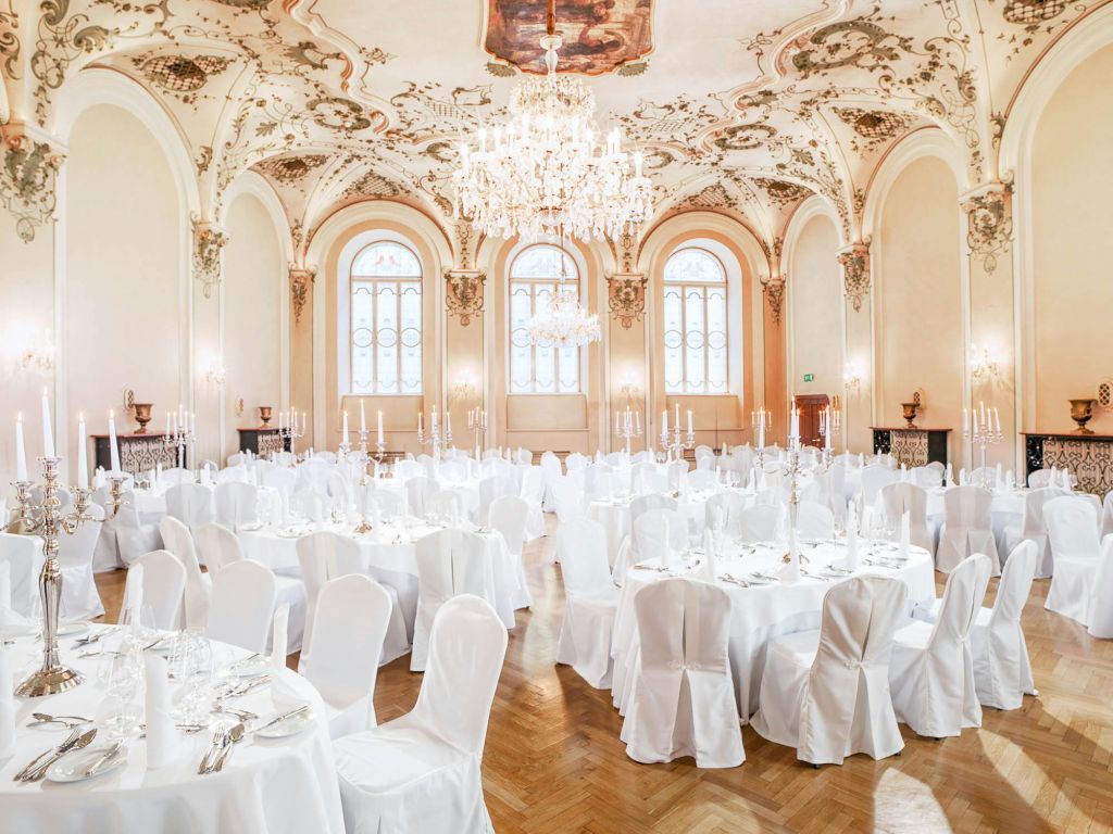 흰색 천으로 덮인 원형 테이블, 흰색 의자 커버, 샹들리에, 화려한 천장 디자인이 있는 행사를 위한 우아한 연회장 세트입니다.