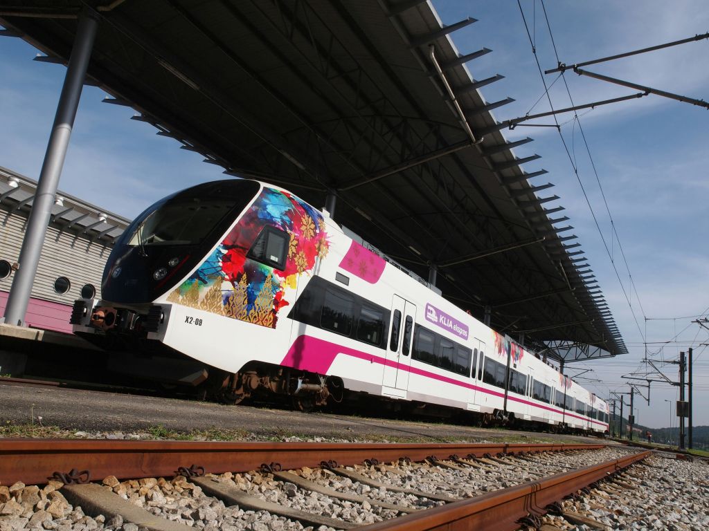 화려한 꽃 디자인과 "KLIA Ekspres" 표지판이 있는 흰색 열차는 기차역의 금속 캐노피 아래에 고정되어 있습니다.