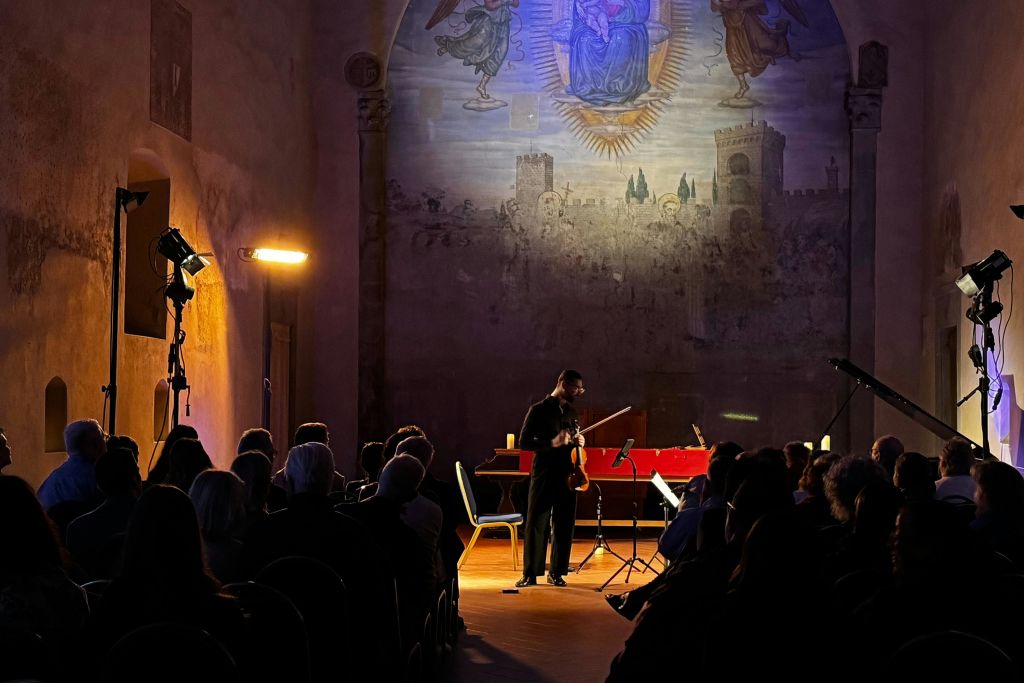 바이올리니스트가 벽에 걸린 커다란 벽화가 관객에게 둘러싸인 희미한 조명으로 장식된 홀에서 공연을 펼치고 있습니다.