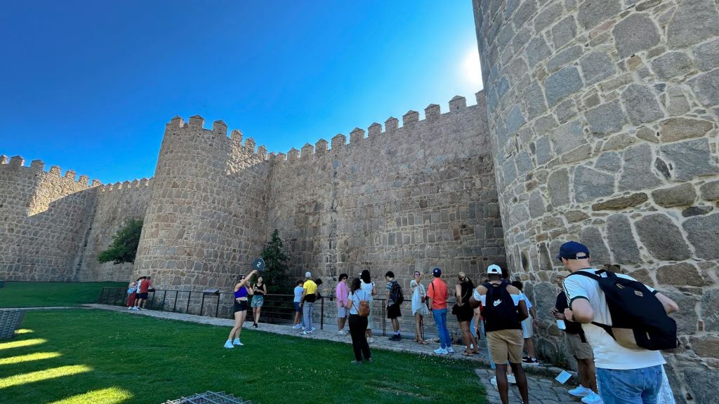 한 무리의 사람들이 맑고 푸른 하늘 아래, 크리넬레이션이 있는 커다란 석조 성벽 근처에 서 있습니다.