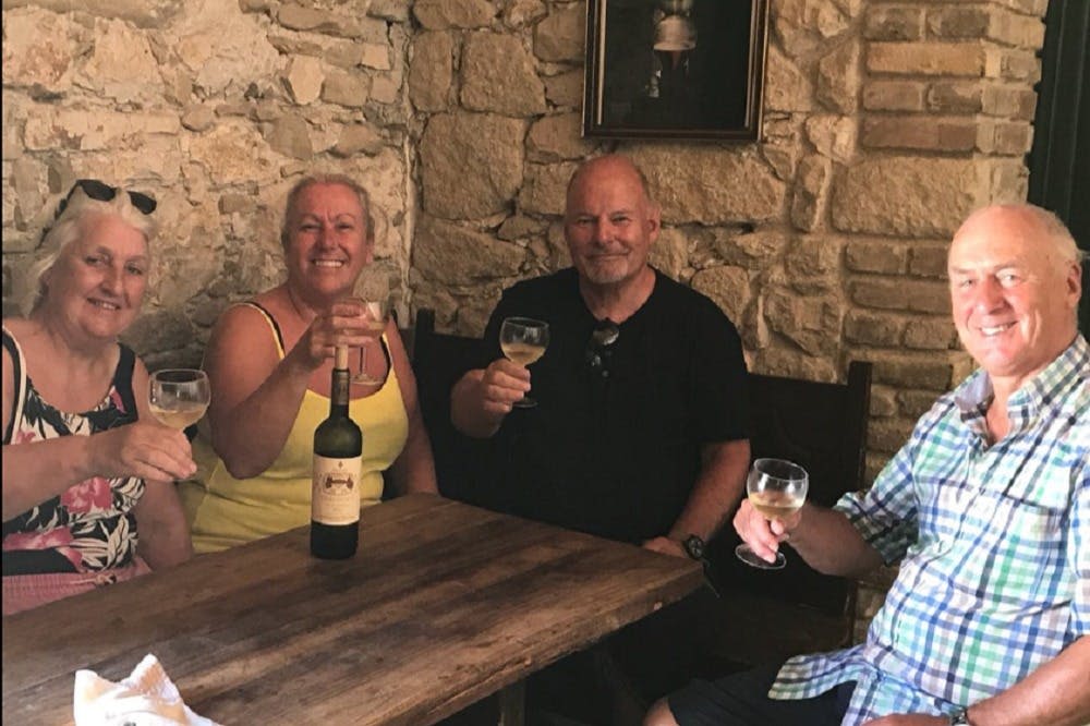 나무 테이블에 앉아 와인잔을 들고 웃고 있는 세 사람, 돌담을 배경으로 한 사람.