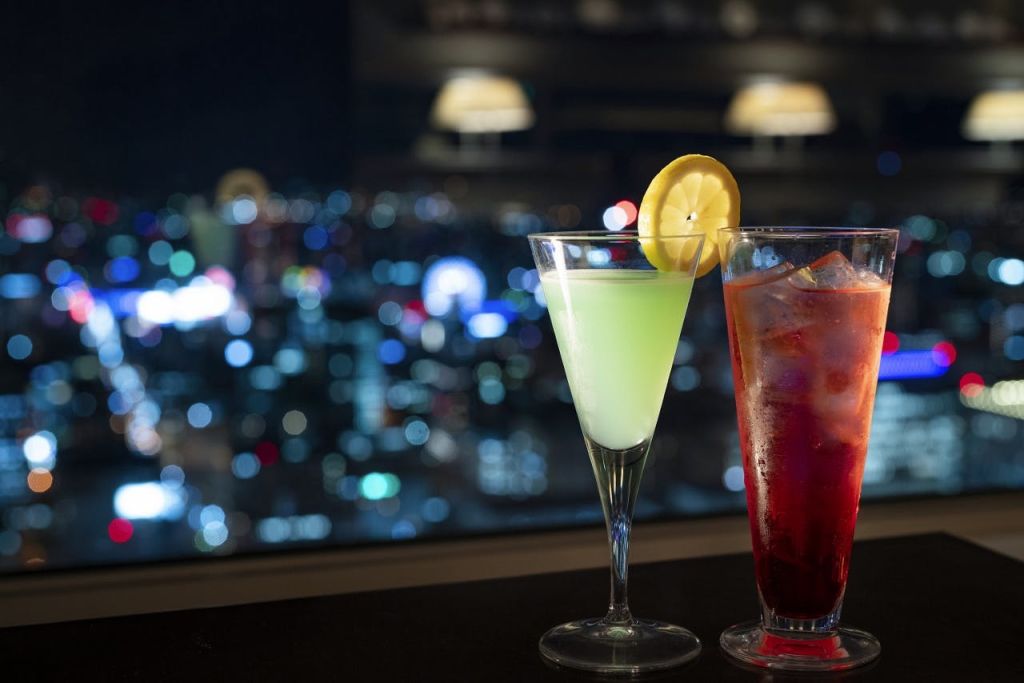 레몬 슬라이스를 곁들인 그린 드링크와 얼음 조각을 곁들인 레드 드링크, 도시 풍경을 배경으로 한 두 가지 칵테일.