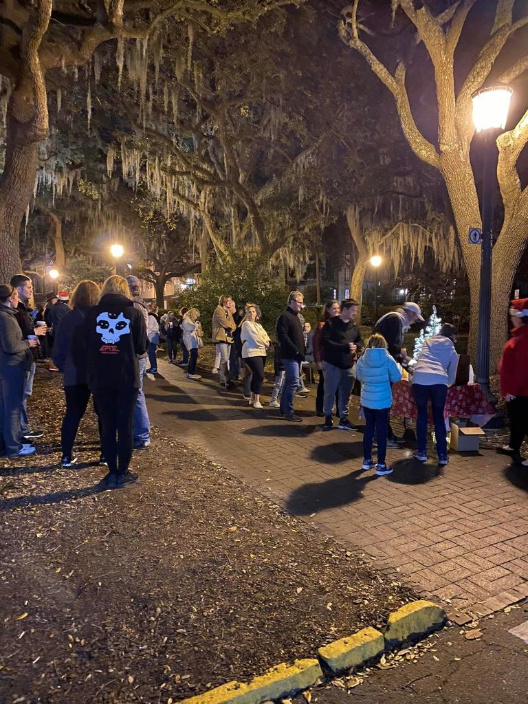 밤에 불이 켜진 가로등 아래 이끼로 덮인 나무와 장식이 놓인 작은 테이블이 있는 공원에 사람들이 모였습니다.