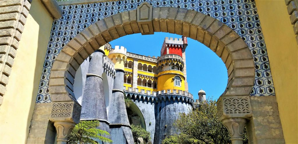 맑고 푸른 하늘 아래, 장식용 석조 아치를 통해 바라보는 노란색, 빨간색, 회색 탑이 있는 다채로운 성.