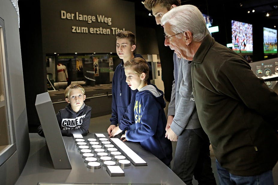 한 무리의 소년과 한 노인이 버튼이 있는 대화형 디스플레이를 살펴보고 있습니다. 벽에 있는 독일어 텍스트에는 "Der lange Weg zum ersten Titel" 이라고 쓰여 있습니다.