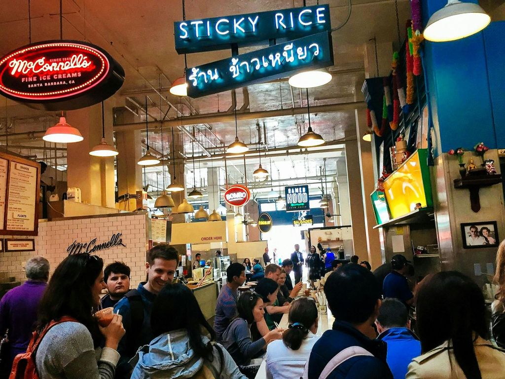 사람들이 식사를 하고 음식을 주문하는 바쁜 실내 시장. 네온 "찹쌀" 과 "맥코넬 아이스크림" 표지판이 보입니다.