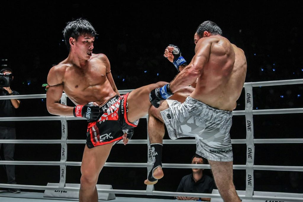 한 반지에 두 명의 무술가가 섞여 있고, 한 무술가는 사진작가를 배경으로 다른 무술가의 미드섹션에 킥킥거림을 전달합니다.