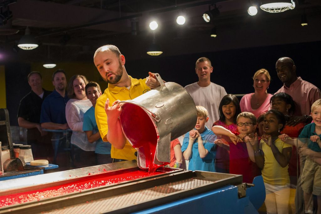 노란색 셔츠를 입은 사람이 큰 용기에서 빨간색 액체를 붓고 어린이와 어른이 신나게 구경합니다.