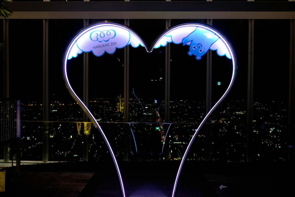 "HARUKAS 300" 텍스트가 있는 하트 모양의 프레임, 만화 곰, 유리창을 배경으로 보이는 도시의 불빛.