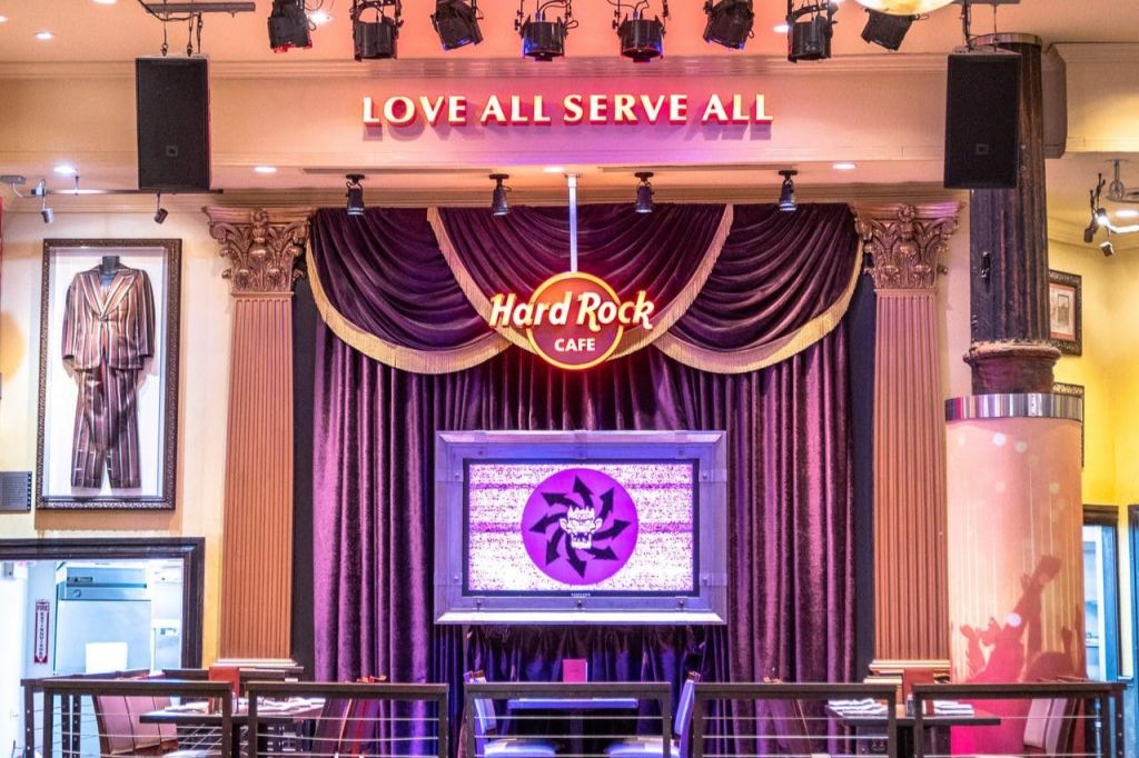 보라색 커튼, 'Hard Rock Cafe' 라고 적힌 로고, 위의 'Love All Serve All' 문구가 있는 무대.