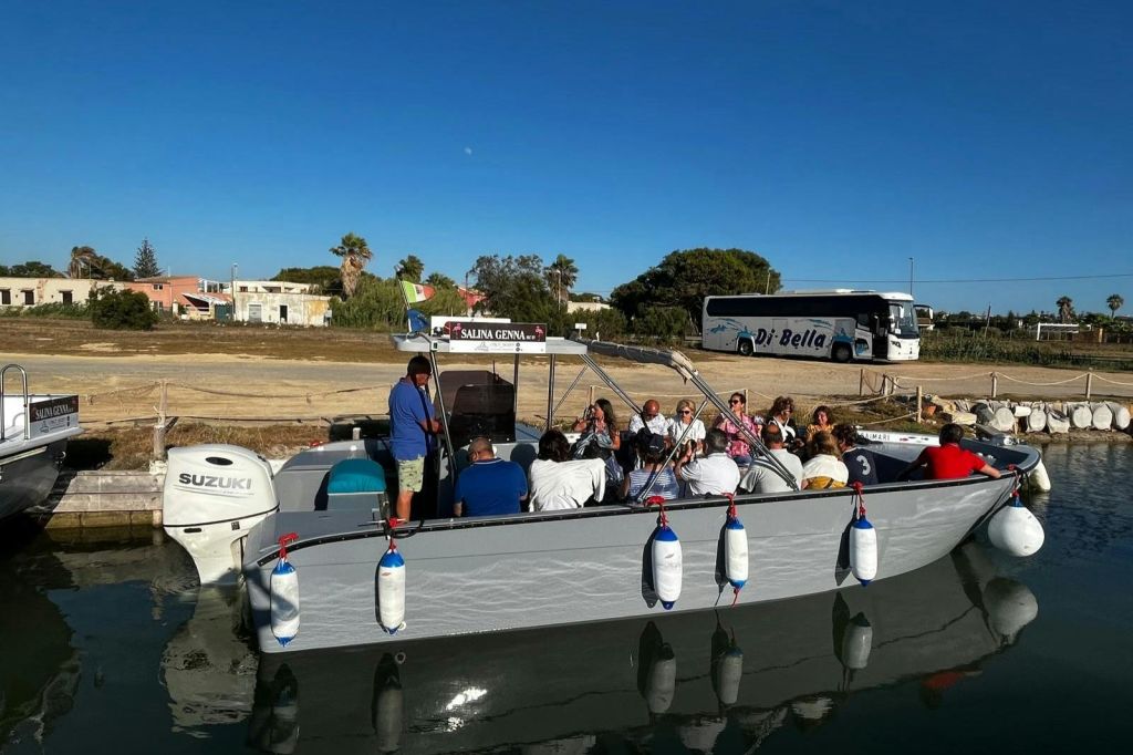 한 무리의 사람들이 운하가 정박한 작은 보트에 앉아 있으며, 맑고 푸른 하늘 아래 배경에 버스와 나무가 보입니다.