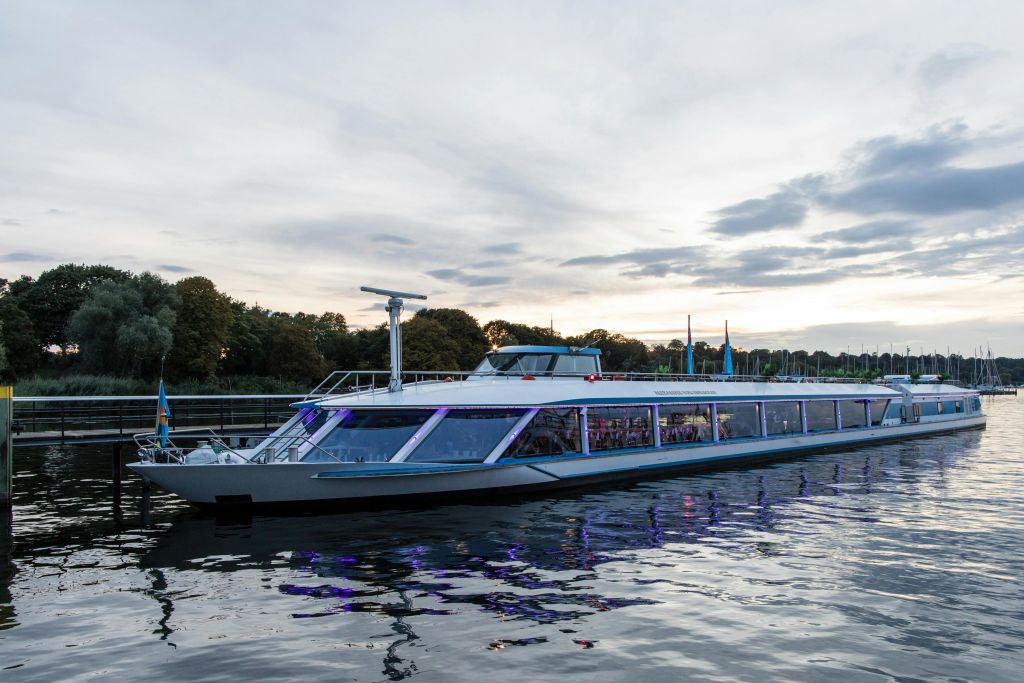 석양이 질 무렵 나무가 늘어선 해안 근처의 잔잔한 물가에 정박한 현대적인 유리벽 보트가 물 속을 비추고 있습니다.