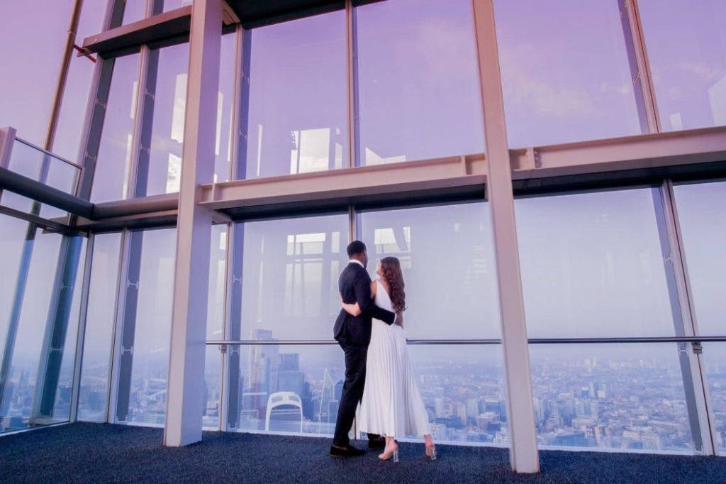 정장 차림의 커플이 도시의 스카이라인 전망을 감상할 수 있는 커다란 창문을 바라보며 보라색 조명 천장 아래 포옹하고 있습니다.