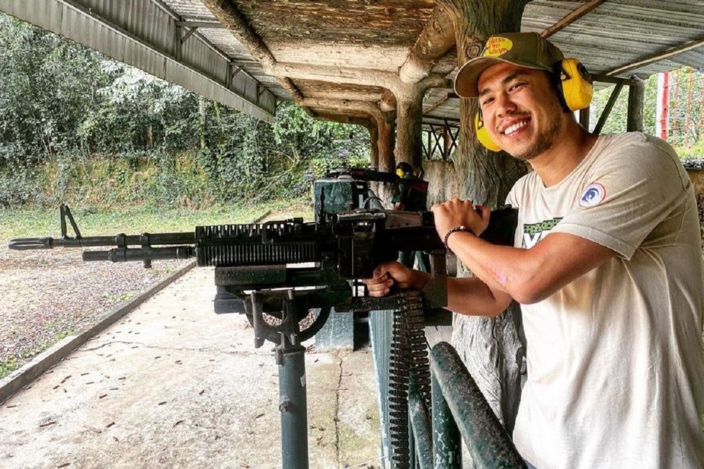 귀를 보호하고 모자를 쓴 웃는 남성이 실외에 설치된 기관총 뒤에 서 있습니다.