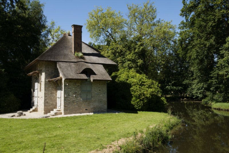 화창한 날 나무와 녹지로 둘러싸인 강 옆에 초가 지붕이 있는 작은 돌집.