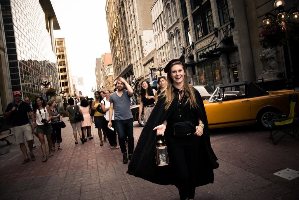 검은 망토와 모자를 쓴 웃는 여성이 한 무리의 사람들과 함께 분주한 도시 거리를 걷는 동안 등불을 들고 있습니다.