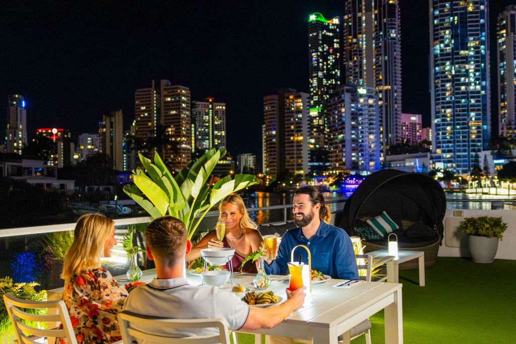 고층 빌딩과 도시의 불빛으로 둘러싸인 야간 야외에서 식사하는 세 사람. 식음료 접시가 테이블 위에 놓여 있습니다.
