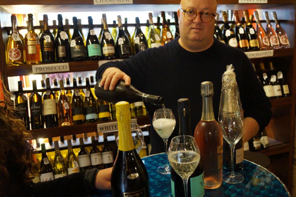 한 남성이 상점에서 다양한 와인 병으로 둘러싸인 테이블 위의 잔에 샴페인을 붓고 있습니다.