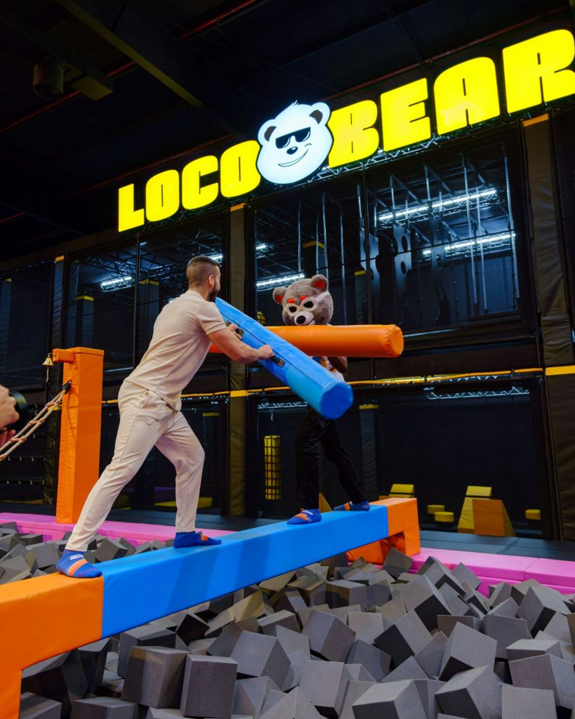 곰 의상을 입은 두 사람이 실내 놀이 센터의 거품 구덩이 위 밸런스 빔에 거품 스틱을 달고 있습니다. 표지판에는 "LOCOBEAR" 라고 쓰여 있습니다.