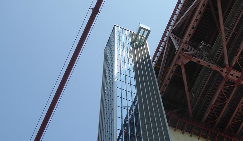 크고 붉은 금속 구조물에 인접한 꼭대기 근처에 돌출된 유리 상자가 있는 높고 유리로 된 건물입니다.